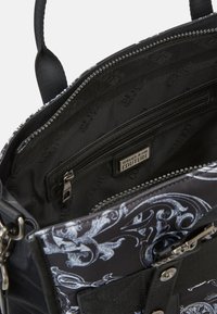 Sort Versace Jeans Couture taske med sølv lynlås, inderlomme med lynlås og hvid barokmønster på stof yderside.