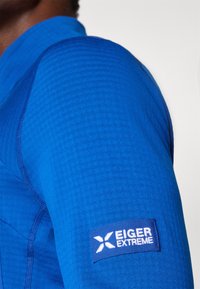 Mėlynas ilgarankovis marškinis su tekstūruota medžiaga, su Eiger Extreme logotipo užrašu ant rankovės. Matomi siūlių detalės.