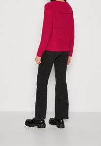 Personne portant un pull en maille rose vif à large col, un jean noir à jambes droites et des chaussures noires à semelles épaisses, debout contre un mur uni.