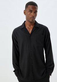 Schwarzes Langarmshirt aus leichtem Stoff mit schmalem Kragen, Brusttasche und Knopfverschluss. Es hat eine lockere Passform und eine weiche Textur.