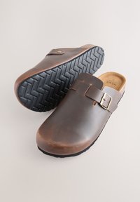 Braune Leder-Clogs mit Schnalle, Kork-Innensohle und einer strukturierten schwarzen Gummisohle mit Zickzackmuster. Glatte Oberfläche.