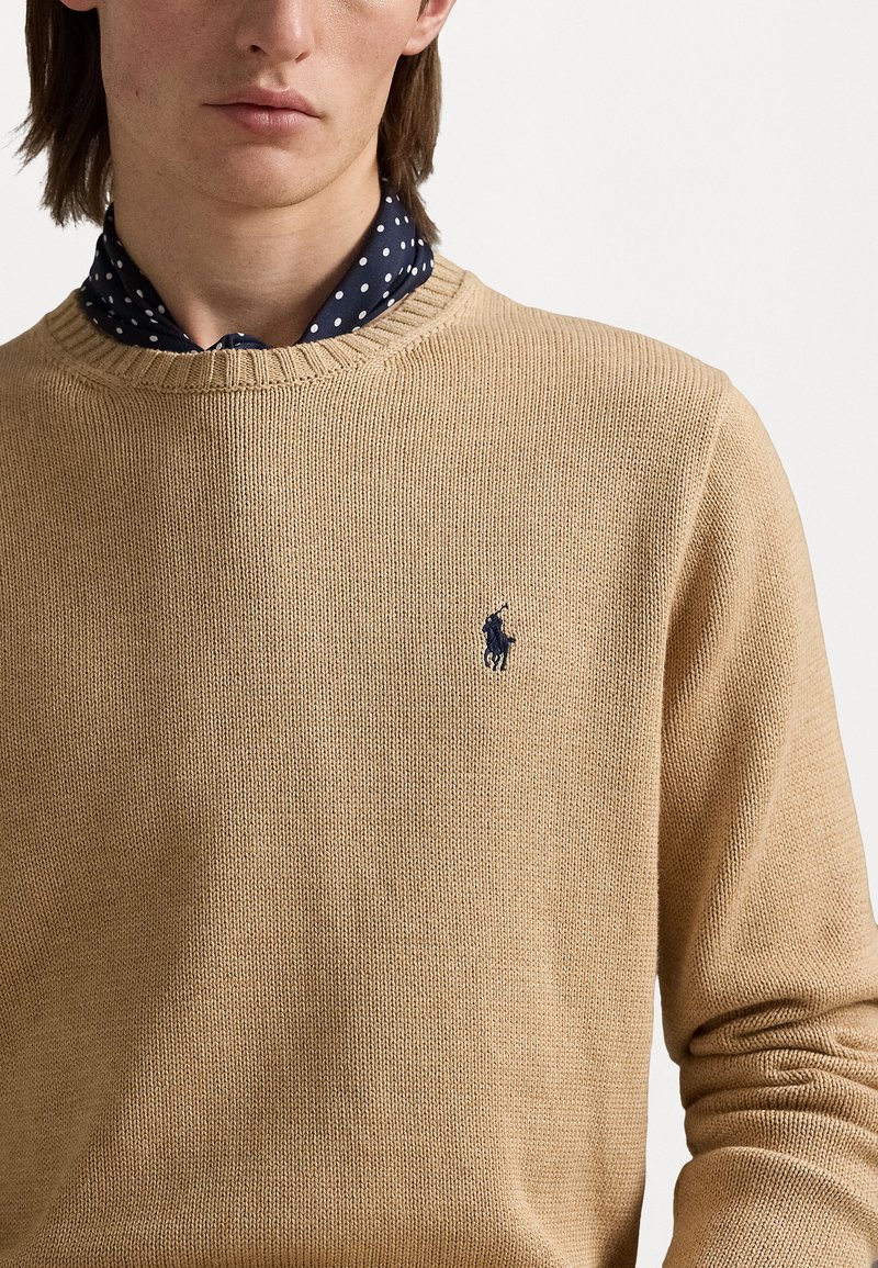 Beige Strickpullover mit Rundhalsausschnitt, wobei das Oberteil ein strukturiertes Muster aufweist und ein kleines Navy-Logo auf der linken Brust trägt.