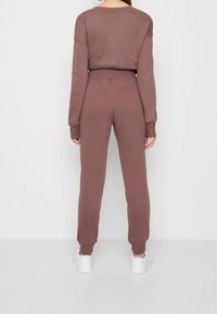 Woman wearing mauve long-sleeve sweatshirt and matching sweatpants, standing facing a white wall, with white sneakers.