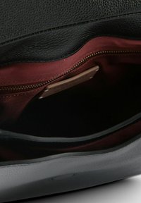 Innenraum einer schwarzen Lederhandtasche mit bordeauxrotem Stofffutter, ausgestattet mit einem Reißverschlusstasche und einem kleinen Etikett, das innen sichtbar ist.