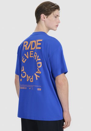 Młody mężczyzna w niebieskiej koszulce z pomarańczowym napisem "RIDE EVERYDAY" ułożonym w okrągły wzór na plecach, stojący bokiem.