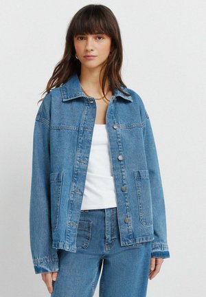 Młoda kobieta ubrana w jasnoniebieską kurtkę dżinsową i dżinsy oraz biały top, stojąca na tle gładkiego jasnego tła, patrząca w kamerę.