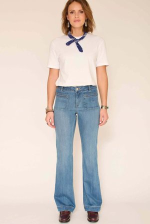 T-shirt blanc, bandana bleu noué autour du cou, jean évasé bleu clair taille haute avec deux poches plaquées, chaussures marron foncé.