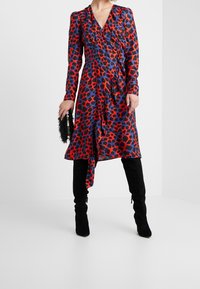 Långärmad omlotklänning i röd och blå leopardmönster, med rynkad design och asymmetrisk nederkant, kombinerad med svarta knähöga stövlar.