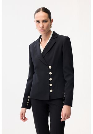 Kvinde med bagudglattet hår iført en tætsiddende sort blazer med fem karakteristiske sølvknapper og matchende sorte bukser.