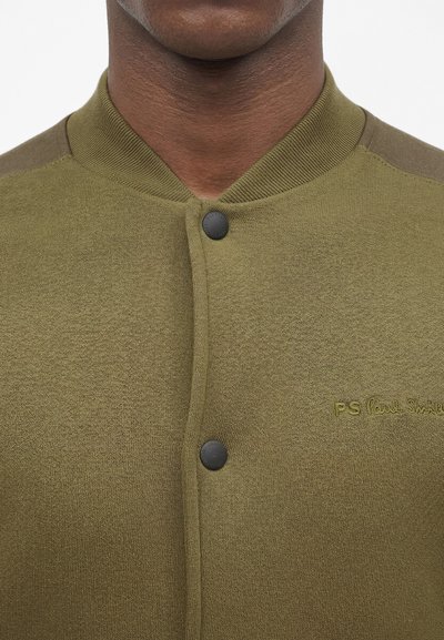 Κοντινή λήψη ενός πλεκτού μπουφάν σε ελιά χρώμα με μαύρα κουμπιά τύπου κουμπί και μικρό κεντημένο λογότυπο «PS Paul Smith» στο στήθος.