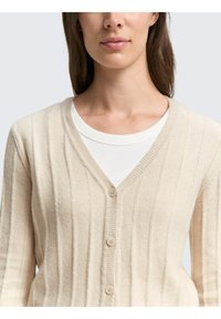 Cardigan bej cu model în dungi, cu decolteu în V, are cinci nasturi și o țesătură texturată, stratificat peste un tricou alb cu guler rotund.
