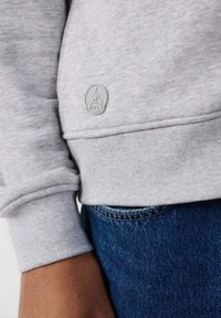 Sudadera gris con puños acanalados, que presenta un pequeño parche de goma gris etiquetado "JOTT" en la parte inferior de la manga, acompañado de vaqueros azules.