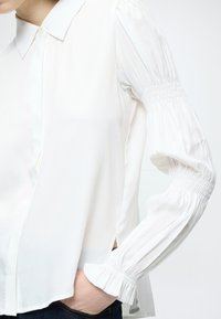 Bílá košile s dlouhým rukávem, límečkem, řasenými rukávy a bočními rozparky. Vyrobena z lehké látky, s jemným nařasením na manžetách.