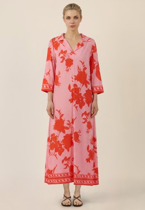 Kobieta ubrana w długą różową sukienkę w czerwone kwiatowe wzory, z rękawami do łokci, kołnierzykiem i sandałami, stojąca na beżowym tle.