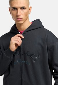 Sort zip-up hoodie med et tekstureret logo; har en hætte med snoretræk og ribbede manchetter. Model iført en kontrasterende pink skjorte under.