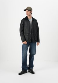 Veste noire matelassée à boutons avec deux poches avant, portée sur une chemise grise. Associée à un jean bleu et des bottes en cuir noires, surmontée d'une casquette à carreaux.