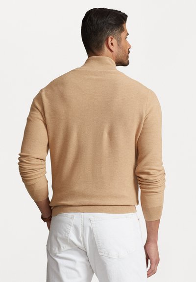 Beige gebreide sweater met een hoge kraag en ribgebreide manchetten. De textuur lijkt fijn en glad, met een getailleerd ontwerp en lange mouwen.