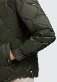 Olijfgroene gewatteerde jas met een geborduurd ontwerp, zijzakken en logotags aan de onderkant en nabij de voorkant.