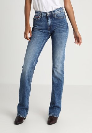 Femme portant un jean bleu délavé droit, une chemise blanche rentrée et des chaussures à bouts pointus marron foncé, debout devant un fond uni.
