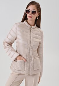 Veste beige rembourrée avec bordure texturée, fermeture éclair à l'avant et poches frontales. Matière lisse avec éléments de design matelassés.