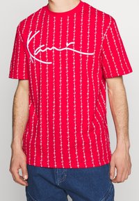 Röd t-shirt med kort ärm, vit ankarmönster och en texturerad, broderad logotyp över bröstet, med en avslappnad passform och rundad halsringning.