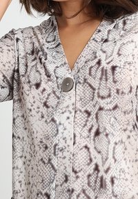 Blouse légère à motifs dans des tons de gris et beige, avec un col en V et un grand bouton à l'avant. Texture subtile à imprimé serpent.