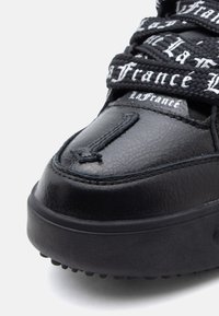 Μαύρο αθλητικό παπούτσι με υφή μπροστά, με χοντρή σόλα και μαύρα κορδόνια που έχουν εκτυπωμένο το "La France" σε λευκή γοτθική γραμματοσειρά.