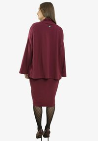 Kobieta w bordowej kurtce z długim rękawem i spódnicy, czarnych rajstopach z wzorem oraz bordowych szpilkach, stojąca tyłem do widza.