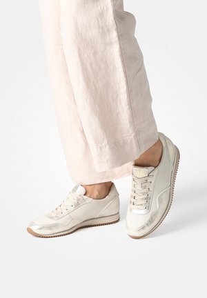 Light beige sneakers with a glossy finish, lace-up design, and beige rubber sole. Paired with light, loose-fitting pants.