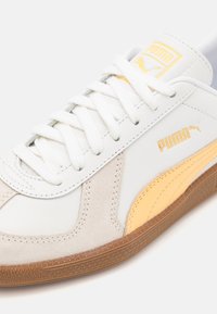 Scarpa da ginnastica bianca con dettagli gialli e in suede grigio. Presenta un design con lacci, suola in gomma e marchio Puma in oro.