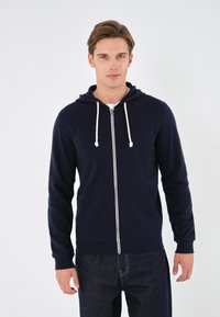 Mornarska zip-up dukserica od mekanog pamuka, s prednjim patentnim zatvaračem, kapuljačom s vezicom i dva prednja džepa. Riblji manžeti i donji rub.