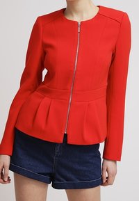 Femme portant une veste rouge ajustée à fermeture éclair avec des plis à la taille et un short en denim bleu foncé, se tenant devant un fond clair.