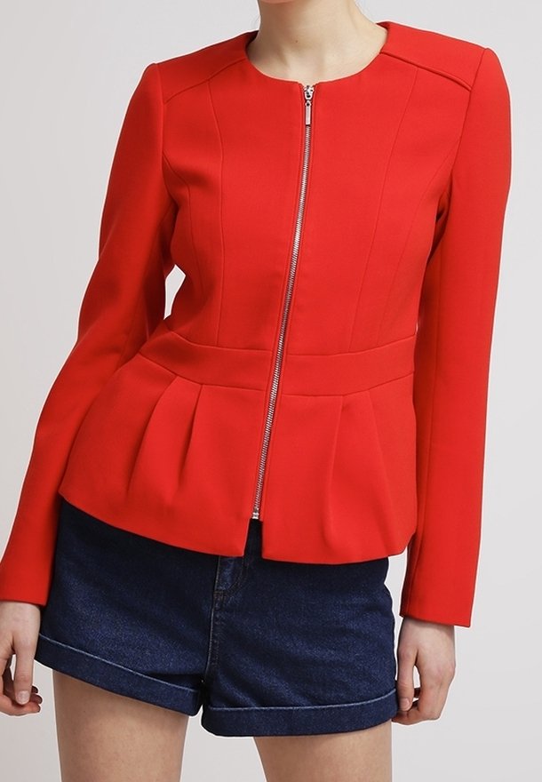 Femme portant une veste rouge ajustée à fermeture éclair avec des plis à la taille et un short en denim bleu foncé, se tenant devant un fond clair.
