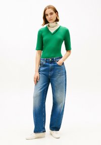 Top verde a righe con maniche corte abbinato a jeans azzurri a taglio comodo. Il modello indossa una sciarpa leggera e sneakers bianche. Look semplice e casual.