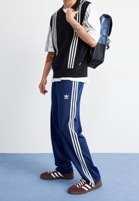 Svart ärmlös tröja med vita ränder över en vit t-shirt, ihop med marinblå träningsbyxor med tre vita ränder och Adidas-logotyp.