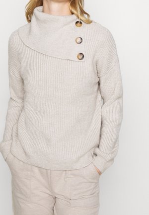 Beige strikket sweater med asymmetrisk fold-over krave, fastgjort med tre store brune knapper, kombineret med lysebeige bukser.