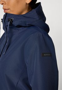 Marineblå jakke med høy krage og justerbar snorhette, med en matt tekstur og et logo-patch på venstre erme.