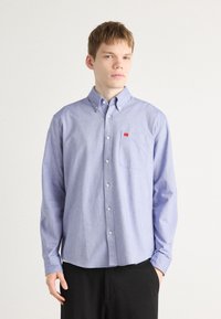 Lyseblå skjorte med knapper og brystlomme, laget av et mykt stoff. Har en liten rød logo på lommen og knappedeler.