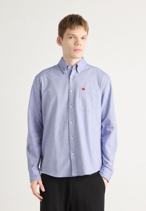 Light blue button-up shirt with a chest pocket, made from a soft fabric. Features a small red logo on the pocket and buttoned cuffs.