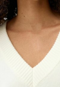 Zbliżenie na szyję i górną część klatki piersiowej osoby noszącej biały, prążkowany, V-kształtny sweter, na szyi widoczne dwie małe znamiona skórne.