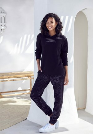 Black sweatshirt with "HAPPY MOMENTS" text, paired with black patterned pants featuring floral designs, and white sneakers. Smooth textures.