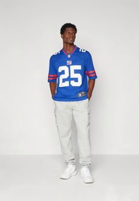 Jersey azul de la NFL con detalles blancos y rojos, número 25. Combinado con pantalones cargo gris claro y zapatillas blancas. Corte sencillo y relajado.