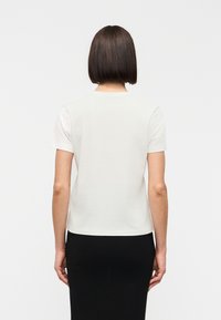 Άτομο με κοντά σκουρόχρωμα μαλλιά που φοράει απλό λευκό κοντομάνικο πουκάμισο και μαύρη φούστα, εμφανιζόμενο από πίσω με φόντο λευκό.