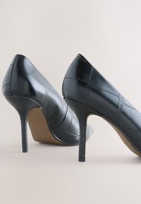 Paire de chaussures à talons noirs avec motif en cuir cousu, vues de dos et de côté sur un fond clair uni.