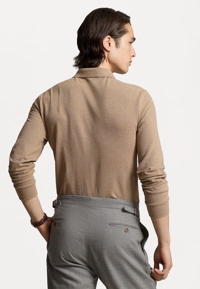Beige poloshirt met lange mouwen van een zachte stof. Grijze op maat gemaakte broek met achterzakken. Het model is van achteren te zien.