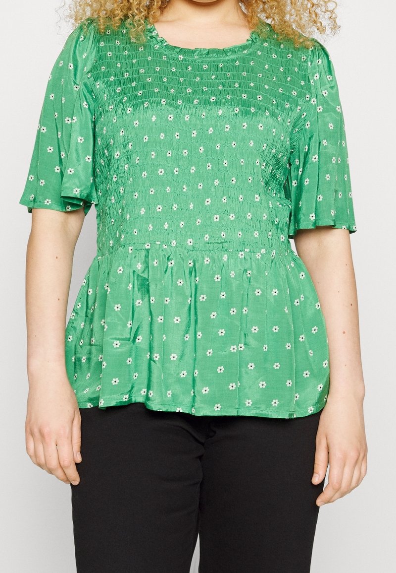 Grøn bluse med blomsterprint lavet af struktureret stof, med korte ærmer, rynkede detaljer og peplum-snit. Båret med sorte bukser.