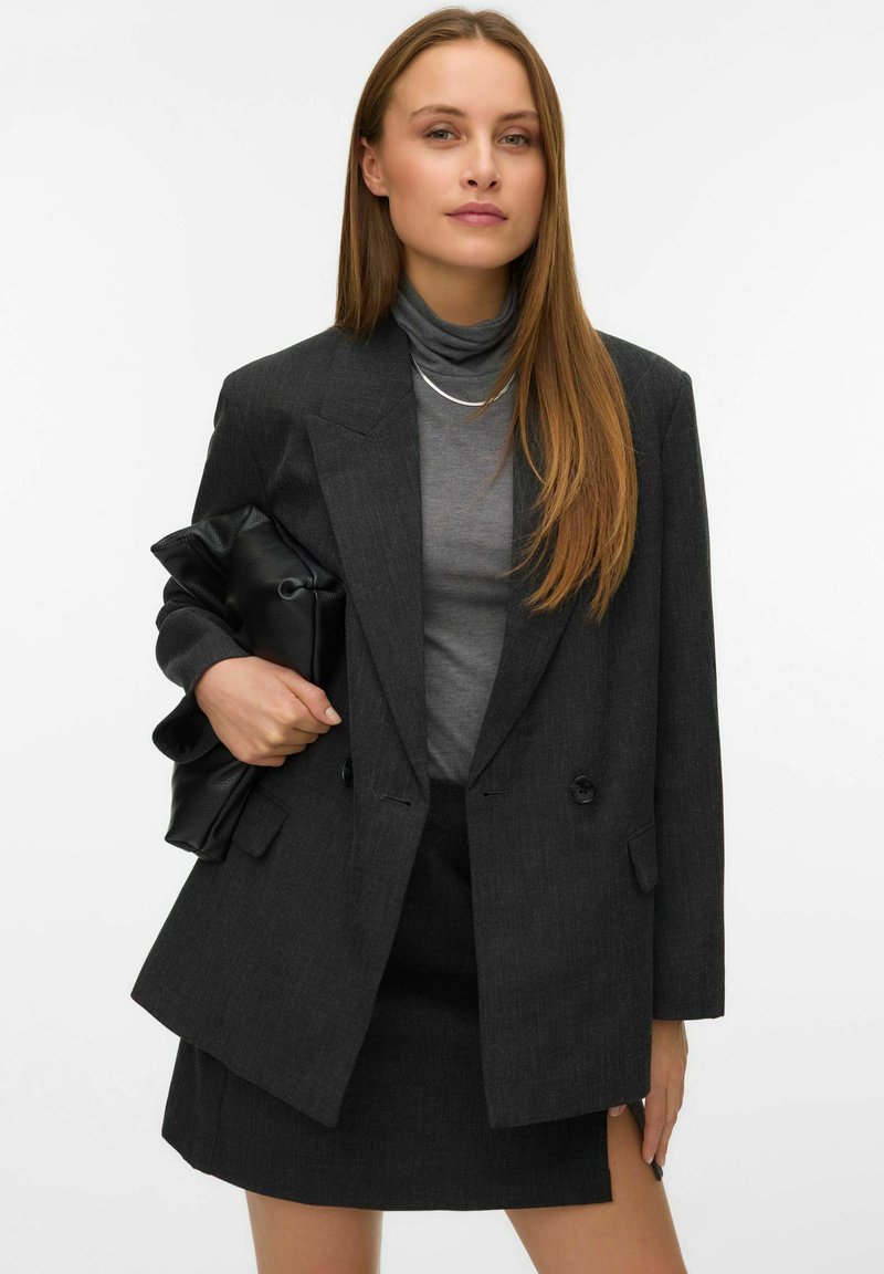 Žena s dlouhými rovnými vlasy má na sobě tmavě šedý blazer a sukni, šedý rolák a drží velkou černou kabelku na pozadí bílé barvy.