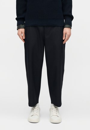 Personne portant un pantalon noir ample, un pull en maille bleu marine avec des poignets noirs, et des baskets blanches, debout devant un fond blanc uni.