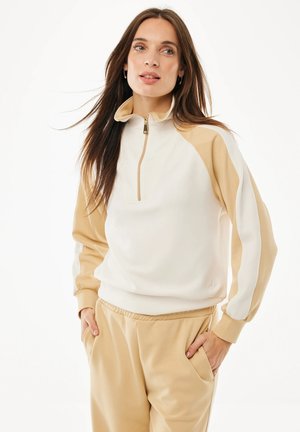 Frau mit langen braunen Haaren, die einen beige-weißen Sweatshirt mit Reißverschluss und beige Hosen trägt, die Hände in den Taschen, schaut leicht zur Seite.