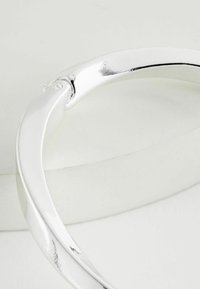 Bracciale in metallo argentato con un design liscio e curvo e una finitura lucida. Presenta un sottile dettaglio attorcigliato vicino alla chiusura.